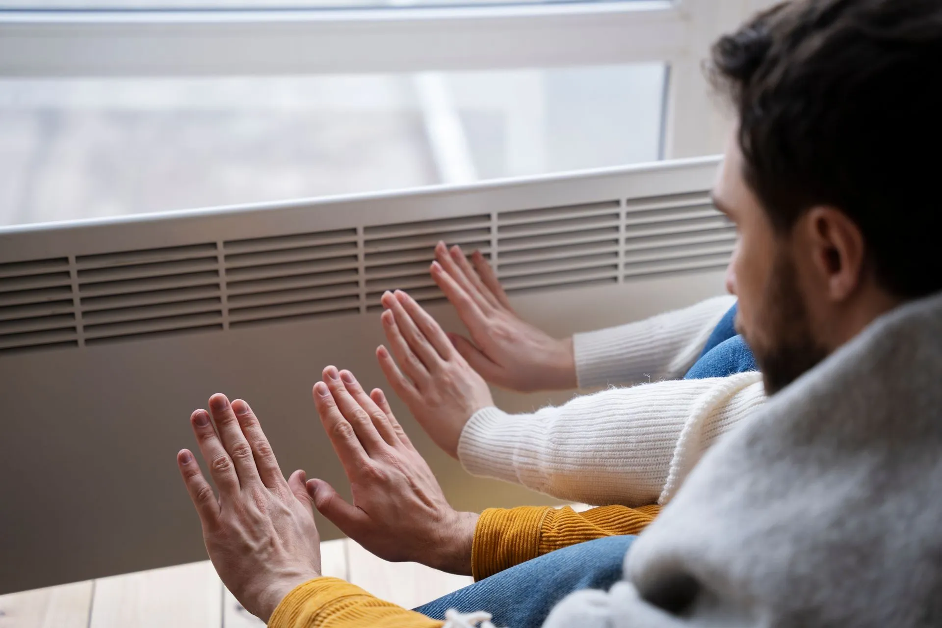 Two people sit side-by-side near a window, holding their hands in front of a wall-mounted heater to warm them.
